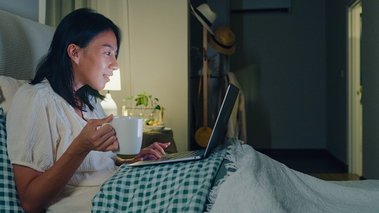 Young Asian girl using laptops and drinking milk sitting on the bed in home at night. Insomnia, Cybersickness, Nomophobia, sleep disorder concept.