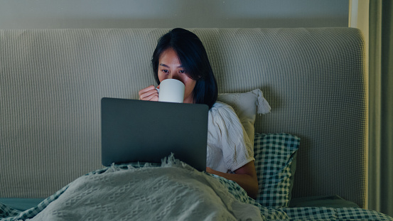 Young Asian girl using laptops and drinking milk sitting on the bed in home at night. Insomnia, Cybersickness, Nomophobia, sleep disorder concept.