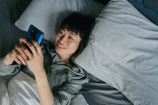 Portrait of a beautiful Japanese woman in her bedroom, still in her pajamas, reading morning news online on her smart phone