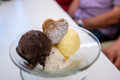 An appetizing cup of ice cream with three yellow, white and beige scoops. A heart-shaped biscuit to garnish it. In the blurred background are the people who ordered it.