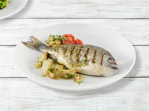 Sea bream fish in service plate with garnish on a white painted wooden table