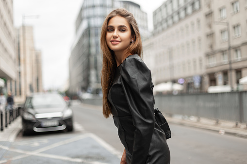 Fashionable happy trendy beautiful girl with smile in black fashionable dress with leather bag walks in the modern city near a road