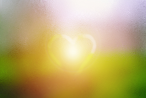 One heart drawn on dewy window glass, fogged with raindrops, sun glare.Selective focus.