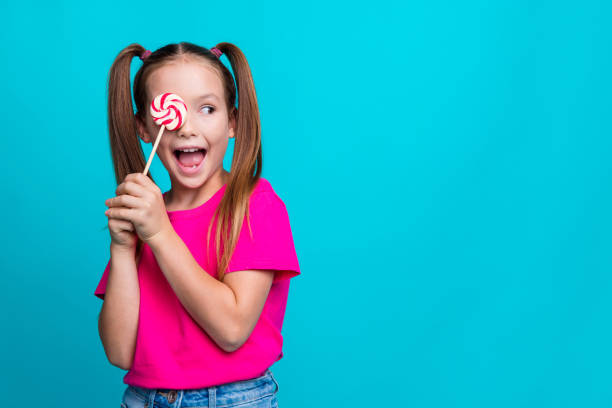 Photo of positive schoolgirl with ponytails wear pink t-shirt hold lollipop on eye staring empty space isolated on teal color background Photo of positive schoolgirl with ponytails wear pink t-shirt hold lollipop on eye staring empty space isolated on teal color background. eye candy stock pictures, royalty-free photos & images