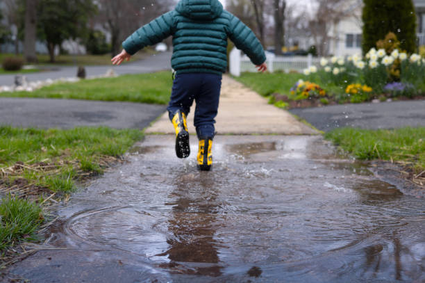 A toddler runs through a puddle in yellow boots with puddle in the foreground A child in yellow rain boots plays and splashes in a puddle after a spring rain april showers may flowers stock pictures, royalty-free photos & images