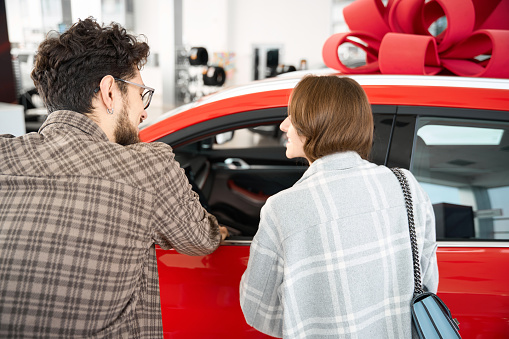 Back view of man and woman standing near car in dealership choosing auto to buy