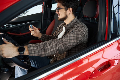 Handsome happy guy holding auto key while sitting in automobile enjoying car purchasing