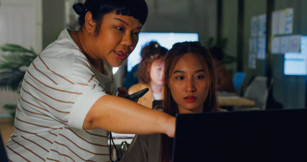 istock Two Asian businesswomen using a smartphone to discuss strategy over a computer in a bustling office environment at night. ESG sustainable business office night. 2119959968