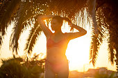 Young woman tying up her hair into bun, while enjoying the sunset under the palm tree on her vacation