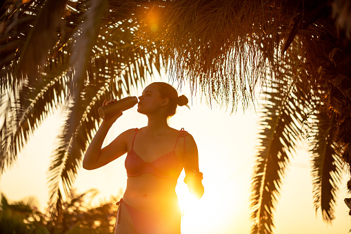Portrait of an young Caucasian woman in swimwear, drinking water, while standing under the palm tree, during sunset