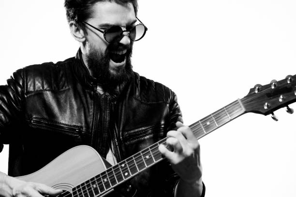 Confidence woman guitarist playing six string acoustic guitar and looking at camera, standing full length. Young adult female musician with glasses is dressed in red leather jacket, jeans and sneakers