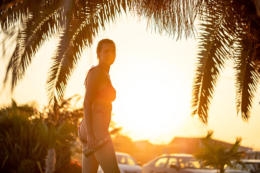 Portrait of an young Caucasian woman in swimwear under palm tree, during sunset