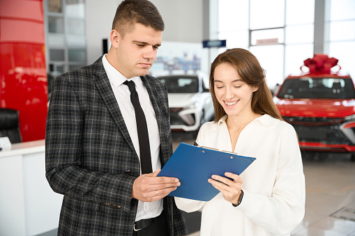Young smiling woman using car rental service signing documents buying automobile in dealership
