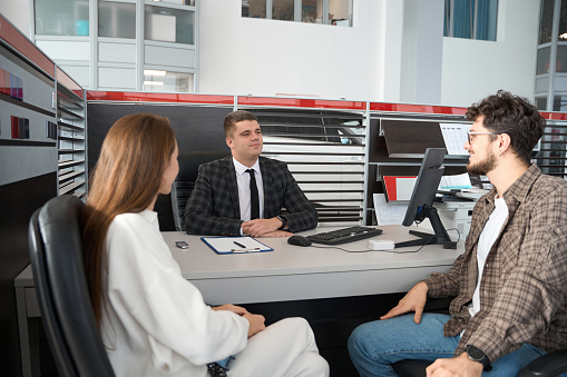 Young couple signing some documents of car purchasing at table with salesperson in dealership office