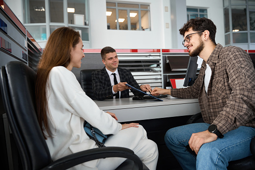 Happy young family talking with dealer customers in automobile salon signing papers in dealership office