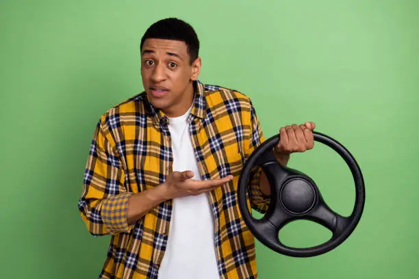 Photo of young man wearing yellow stylish checkered shirt demonstrate volkswagen auto steering wheel isolated on green color background.