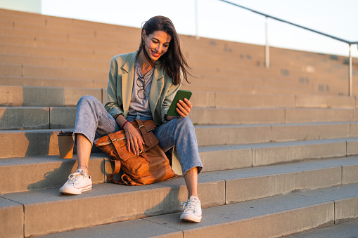 Pretty young smiling woman sitting on steps and reading text messages from friends on smartphone