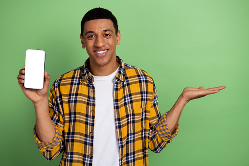 Photo of young cheerful guy wearing checkered shirt hold arm with cellphone promoting digital product isolated on green color background.