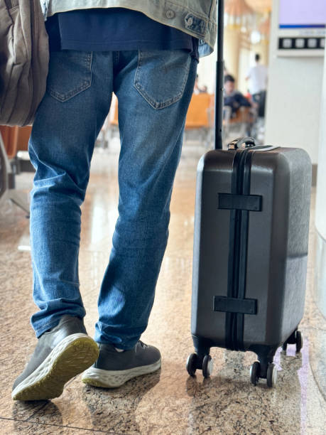 istock Close-up image of unrecognisable passenger wearing denim jacket and shoulder bag, dragging wheelie suitcase carryon luggage behind, airport departure area, shiny tiled flooring, rear view, focus on foreground 2120048301