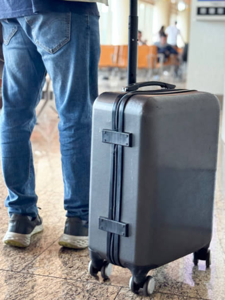 istock Close-up image of unrecognisable passenger wearing denim jacket and shoulder bag, dragging wheelie suitcase carryon luggage behind, airport departure area, shiny tiled flooring, rear view, focus on foreground 2120050335