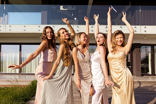 Portrait of excited young women wearing elegant dresses standing in front of villa and raising hands. Female friends during bachelorette party.