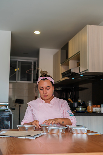 Latina businesswoman from Bogota Colombia between 30 and 34 years old, prepares the packaging where she will send her Italian food to clients wearing her uniform from her kitchen