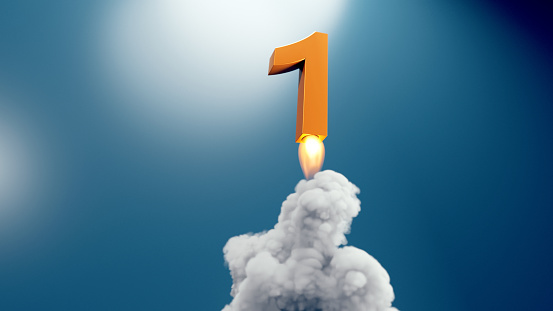 A metallic number one with a bright, ignited flame at its base, resembling the bottom of a rocket during launch. The number flys through the air, with billowing white smoke beneath it, set against a blue background. The scene captures the moment of take off, symbolizing a beginning, achievement or leadership.