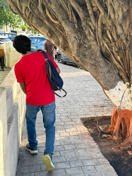 istock Close-up image of man walking on pavement ducking past large ancient Indian banyan (fig) tree trunk painted orange, white stripes forming national flag of India colours with green leaves, aerial roots growing downwards, rear view 2120106403