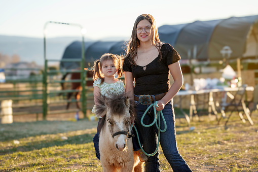 Teenage Girl and Two Year Old Girl Riding Atop a Shetland Pony on a Family Farm at Dusk