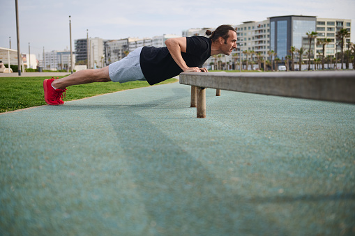 Full length portrait of Caucasian young athletic man doing push ups while working out outdoors. Sportsman pumping up muscles, doing exercises on arms while a bodyweight training in urban environment