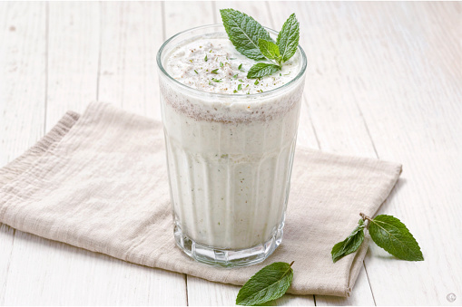 A glass of Turkish traditional drink, ayran, kefir, or buttermilk made from yogurt, garnished with mint leaves, is placed on a beige napkin on a white wooden table.