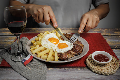 Bistec a lo pobre, beef, eggs, and French fries on a plate on a wooden table with cutlery. Diner seated about to eat, cutting the steak. Wine glass and Chilean flag on the table. Typical Chilean food