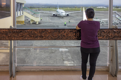 Sultan Iskandar Muda Int'l Airport, Banda Aceh, Sumatra, Indonesia - January 15th 2024:  Woman looking at an approaching Boeing 737 aircraft through an opening in the window film