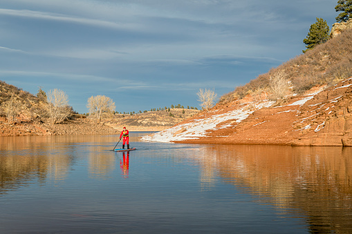 male paddler in red drysuit is paddling a stand up paddleboard on mountain lake in Colorado, winter scenery - self portrait