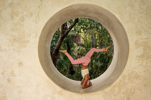 Side view of a young woman performing a yoga pose within a large circular opening in a concrete wall. Dressed in a white sports bra and pink yoga pants, she balances on her head with her legs spread out in opposite direction. Lush tropical foliage can be seen in the background, providing a natural and serene backdrop.