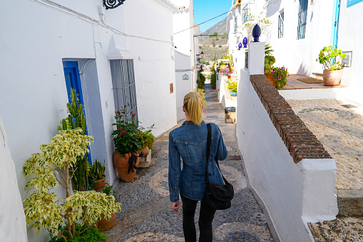 Blond female tourist walking the charming narrow mosaic cobblestone streets and flower pots in the white hillside village of Frigliana in Southern Spain