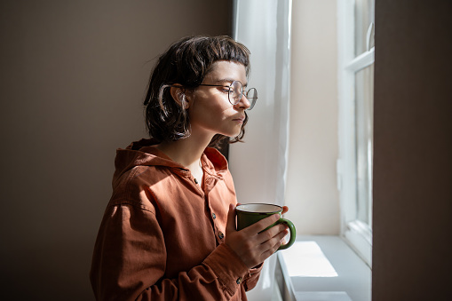 Teen girl nerd in glasses looking at window drinking cup of tea at home. Serious young woman resting contemplating street, watching people cars. Weekend, of introvert, leisure in loneliness concept.