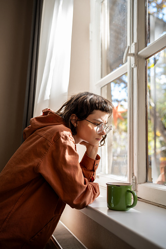 Nerd teen girl in glasses drinking tea looking at window boring at home. Sad upset young woman feeling apathy, age teenage crisis. Mental disorder, anhedonia, stress, having life troubles problems.