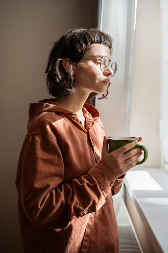 Pensive introvert woman in glasses looking at window drinking cup of tea at home. Girl suffering from mental illness looks outside, watching people, cars, thinking about life problems. Loneliness.