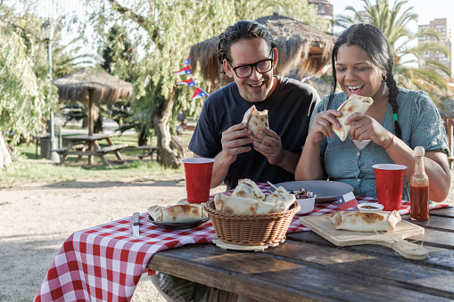 A couple shares an intimate outdoor meal in the park. Both are happy and laughing, about to bite into Chilean pino empanadas. Chilean flag, Independence Day celebrations