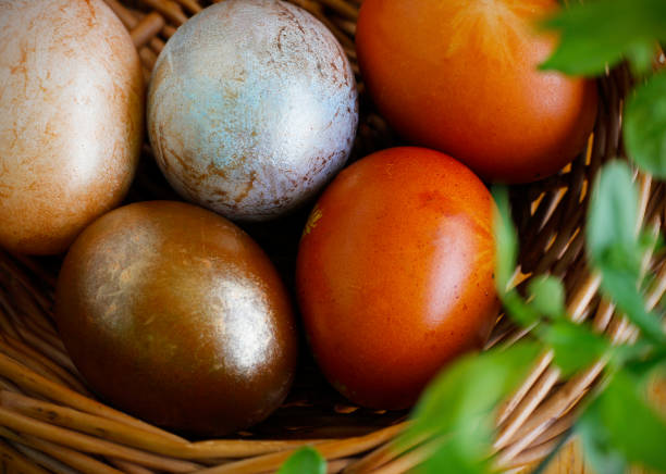 Five Easter eggs painted in different shades of neutral colors lying in a wicker basket under a spring twig with fresh green leaves. Simple Easter flat lay in neutral colors
