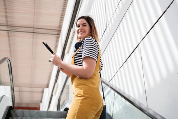 Young woman dressed in yellow dungarees going up the escalator using the tablet. Concept: lifestyle, technology, outdoors.