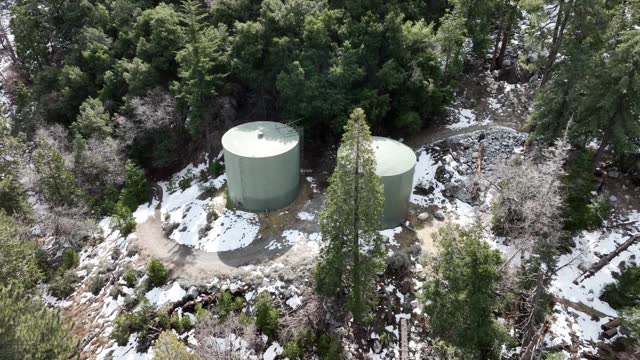 two large water tank reservoirs perched on a snow-covered mountain surrounded with green trees during the day time and a California draught AERIAL ORBIT 60fps