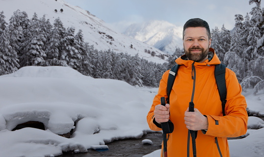 A handsome bearded middle-aged man in an orange traveling jacket holding hiking poles in the forest against the background of a stream and mountains
