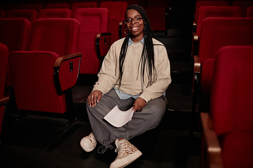 Full length portrait of smiling African American woman sitting on floor in theater and looking at camera copy space