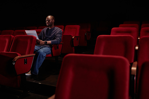 Minimal portrait of African American performer sitting in audience and reading lines in empty theater copy space