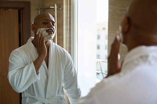 Young african american man smearing shaving foam on face and looking at himself in mirror in bathroom at morning time. Concept of morning procedures and hygiene