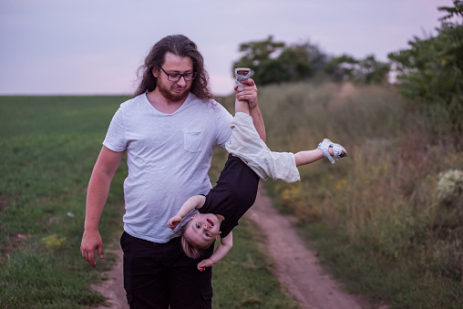 Diversity father holding little son upside down playfully indicates his joyous child. Young man with long curly hair, beard, glasses fooling around with toddler boy on sunset green field