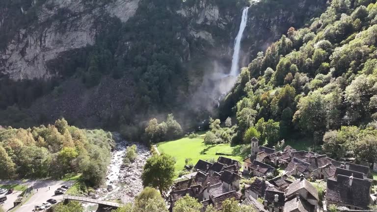 An aerial captures the majestic Foroglio Waterfall in Maggiatal, Tessin, Schweiz. Sunlight illuminates the cascading water against the backdrop of a quaint ancient village nestled in a valley below.