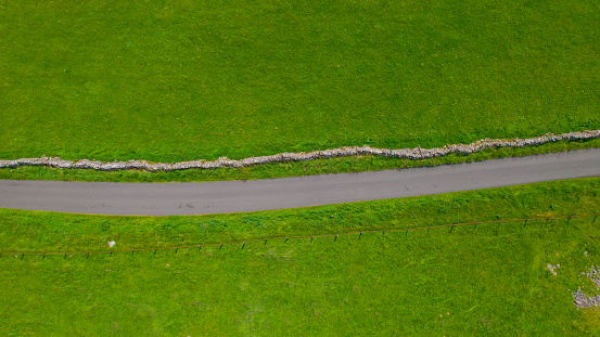 AERIAL TOP DOWN: Asphalt roadway winding past fenced vibrant green pastures, surrounded on one side by a traditional dry stone wall and on the other by a fence. Scenic patterns in English countryside.